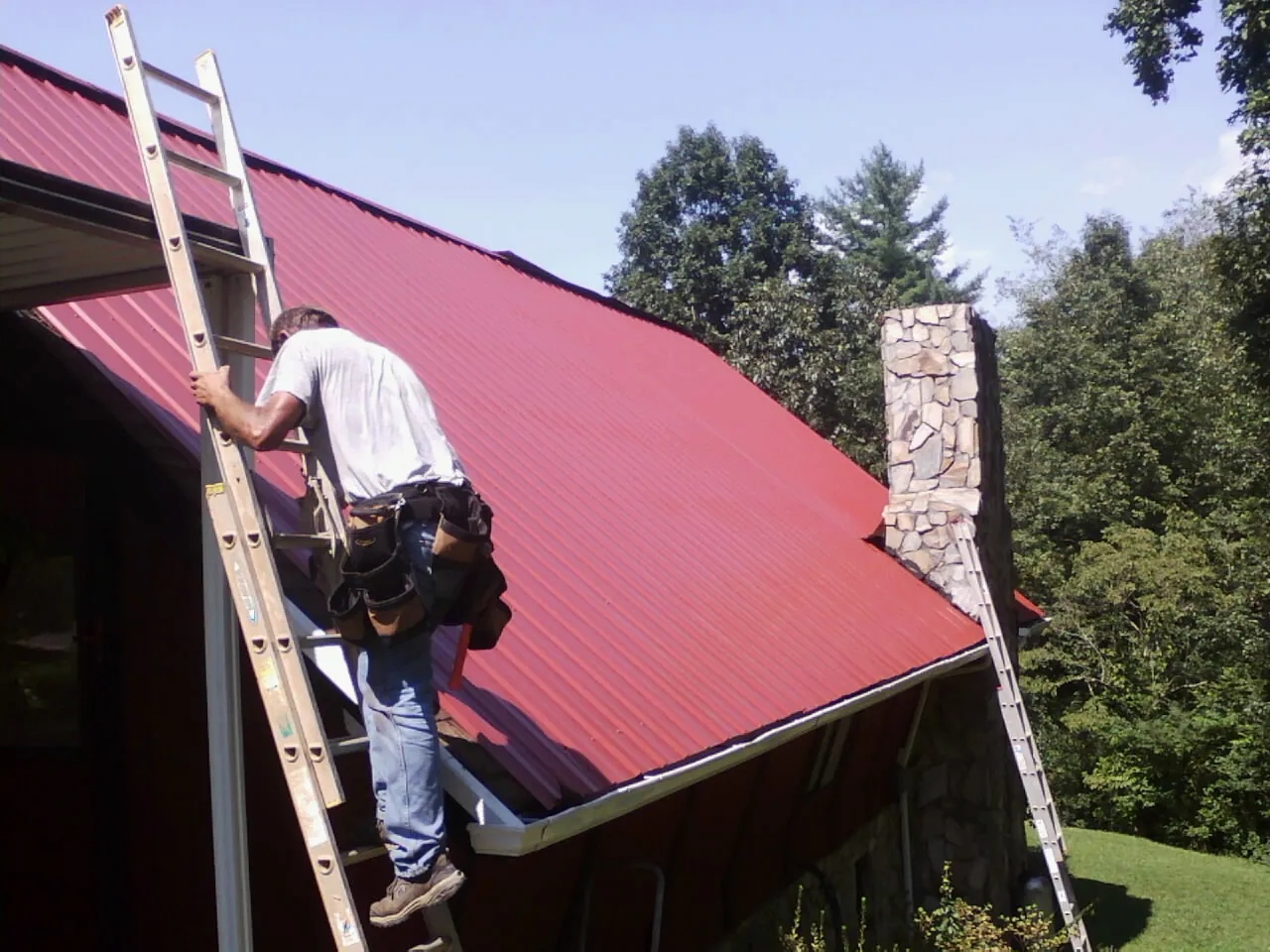 Roofer on a ladder beside a red metal roof and stone chimney — Soffit & Fascia Repair by Roofing Brothers in Lakeside